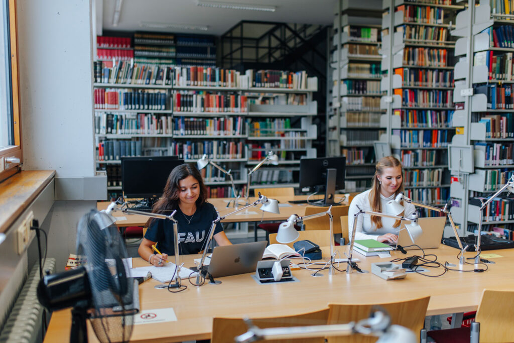 Zwei Frauen sitzen mit ihren Laptops in einer Bibliothek.