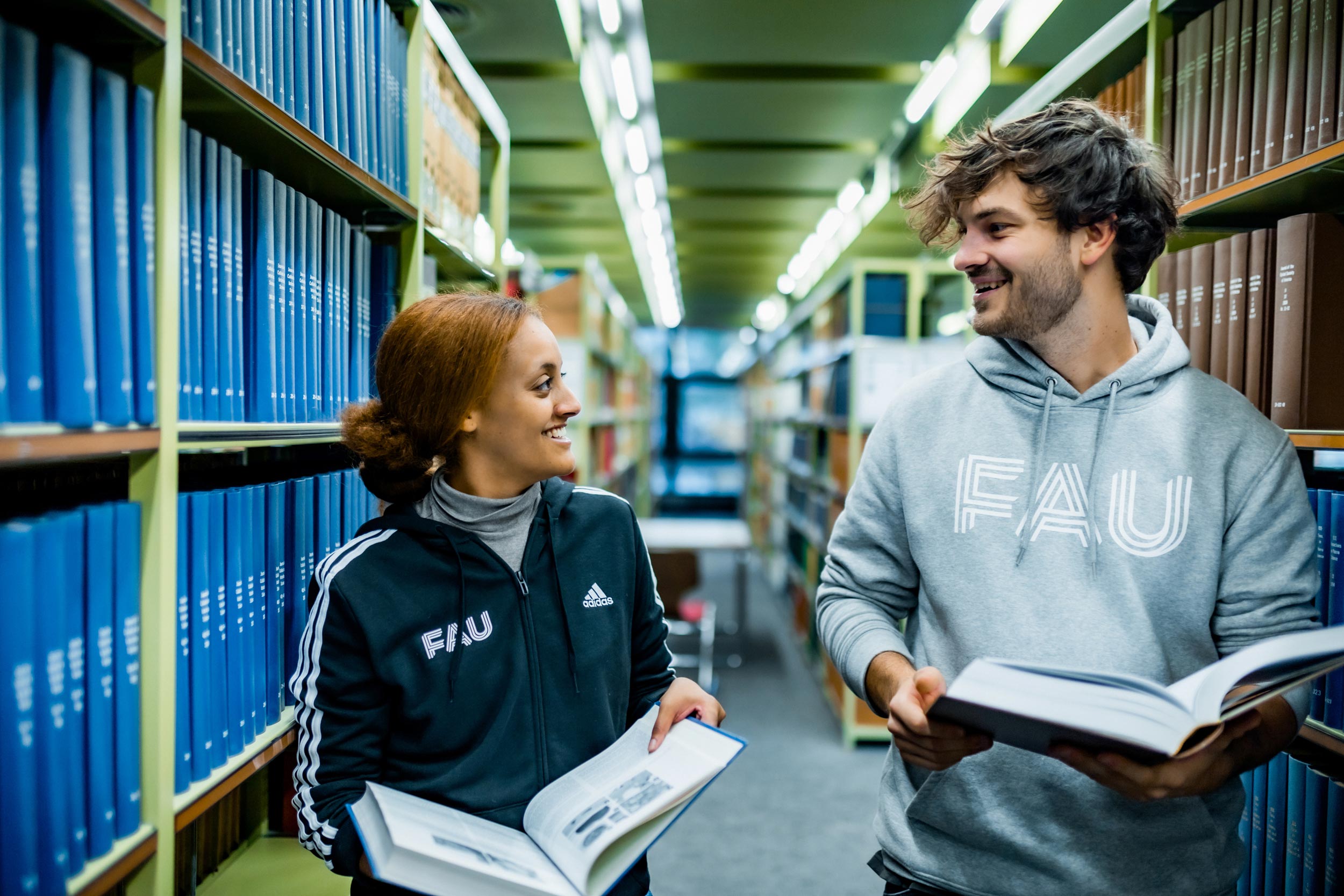 Zwei Studierende lächeln sich an. Sie stehen in einem Gang zwischen Bücherregalen in einer der FAU-Bibliotheken.