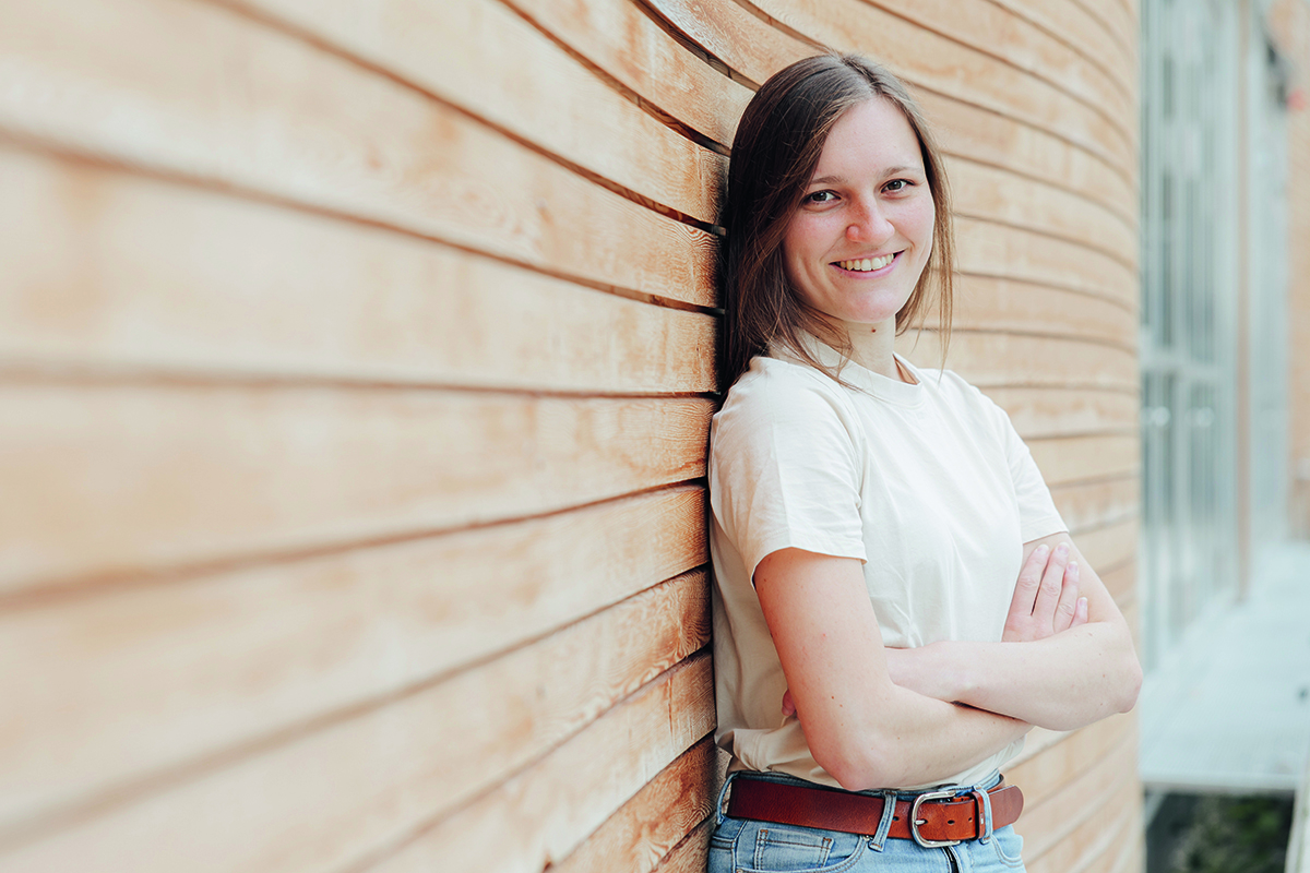 Frau, hein, eine junge frau mit hellbraunem Haar und dunklen Augen, lehnt mit verschränkten Armen an einer Holzwand.