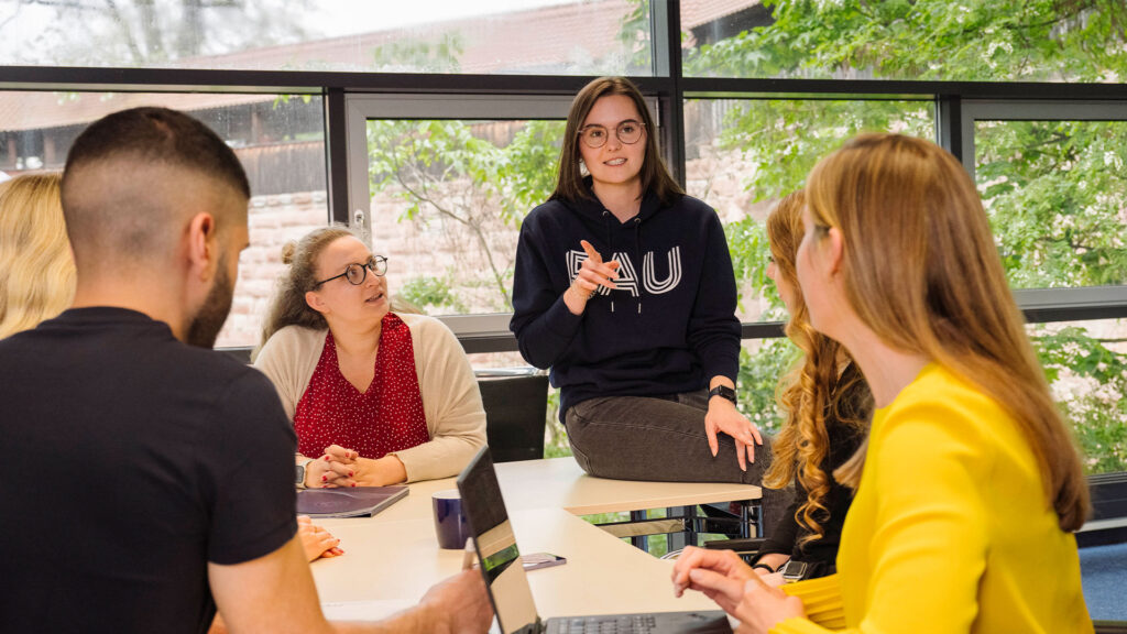 Eine junge Lehrende mit FAU-Pullover sitzt auf einem Tisch und erklärt einer Gruppe etwas.
