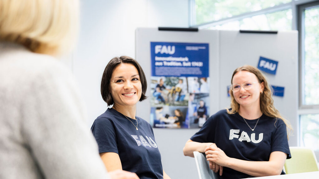 Zwei junge Frauen sprechen mit einer weiteren Person, die von hinten zu sehen ist.