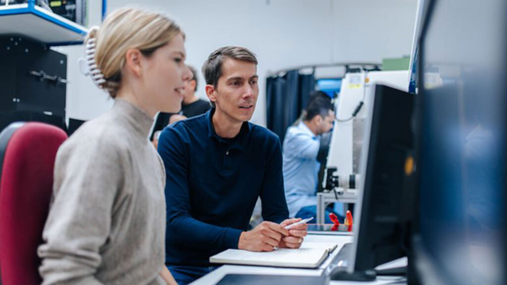 Eine junge Frau und ein Mann sehen gemeinsam auf einen Computermonitor und besprechen sich. Währenddessen machen sie Notizen.
