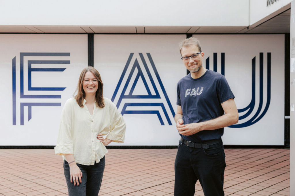 Ein Mann und eine Frau im Außenbereich der FAU vor einer weißen Wand mit FAU-Logo.