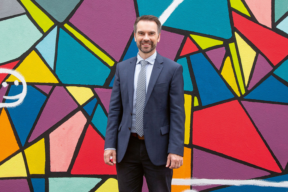 Ramming, ein Mann mit kurzen Haaren und Bart, steht vor einer bunten Wand.