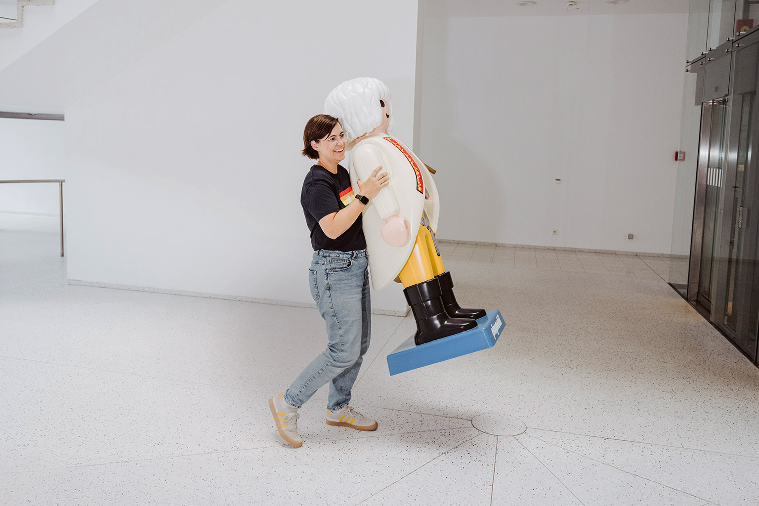 Laura Kneppe, eine Frau mit kurzen dunklen Haaren und Brille, trägt eine übergroße Playmobilfigur.