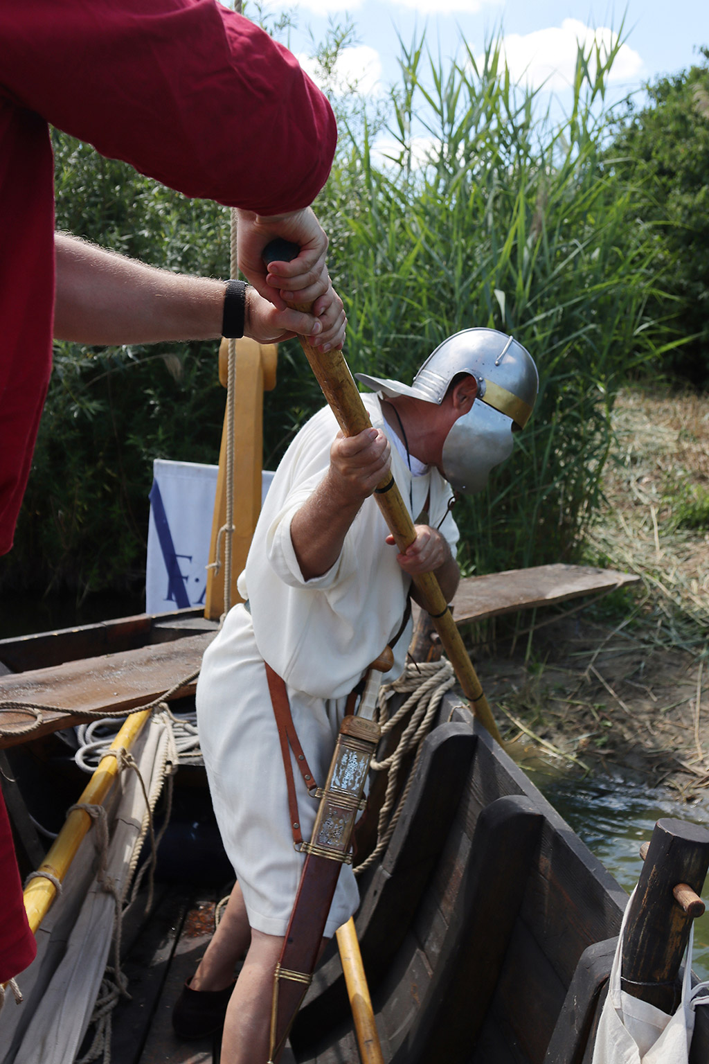 Ein Mann in weißer Tunika mit Helm auf dem Kopf am Ufer eines Sees.