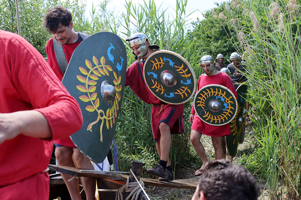 Männer in roten Tunikas und Schilden steigen in ein Boot.