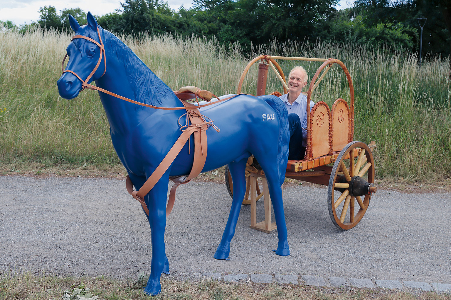 Boris Dreyer sitzt lachend auf einem hölzernen Wagen vor den ein blaues Plastikpferd gespannt wurde.