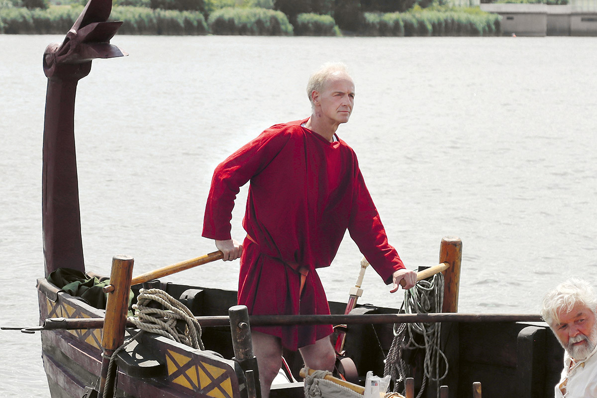 Boris Dreyer in roter Tunika an Bord eines römischen Bootes. Er steht am Steuerruder.
