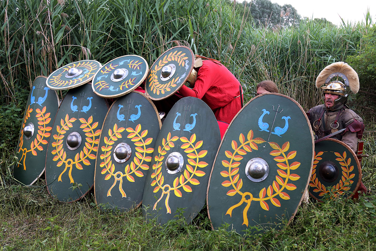 Mehrere Männer in roten Tunikas und römischen Soldatenuniformen verstecken sich hinter einem Schildwall.