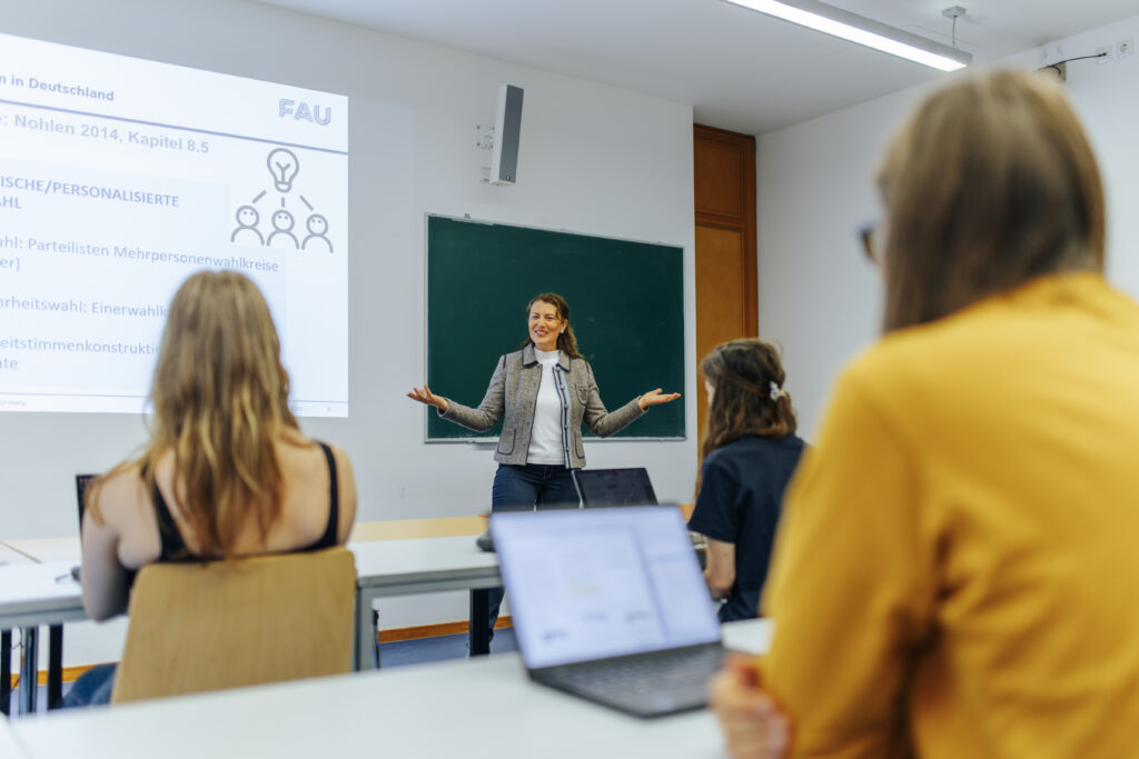 Dozentin zeigt eine Präsenation, drei Personen hören zu.