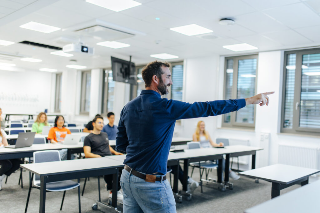 Ein Dozent zeigt auf die Tafel, im Hintergrund sitzen Studierende