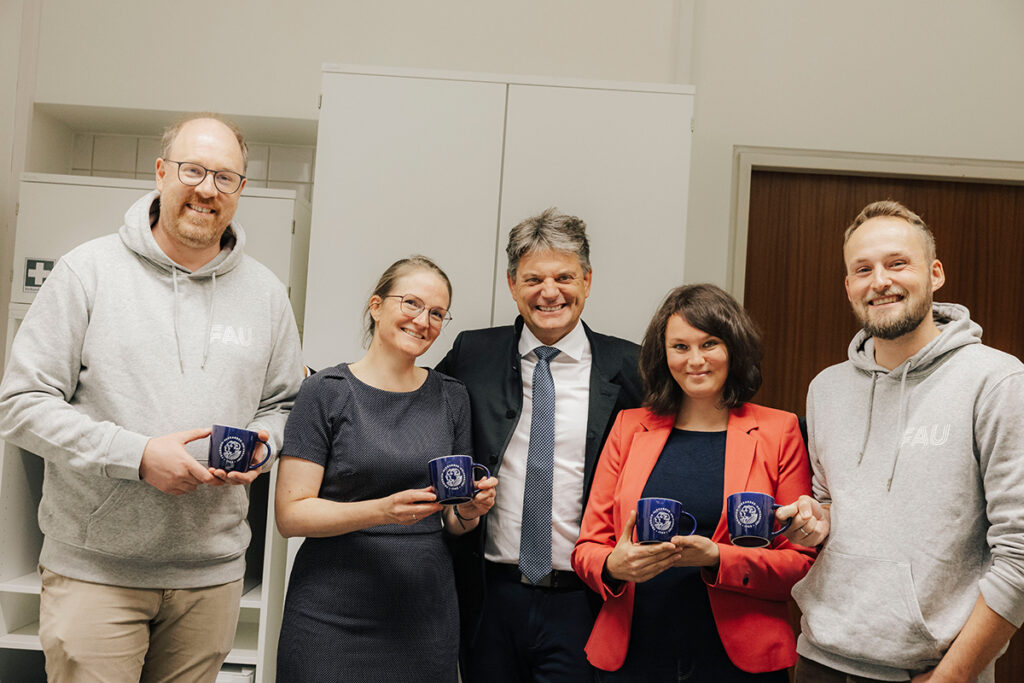 Ein Gruppenbild mit vier Professoren und Professorinnen und dem Präsidenten der FAU. Professor Hasselhorn und Professorin Lindmeier stehen links, Präsident Hornegger mittig und Professorin Berner und Professor Habig stehen rechts. Alle lachen in die Kamera, die Professoren halten FAU-TAssen.