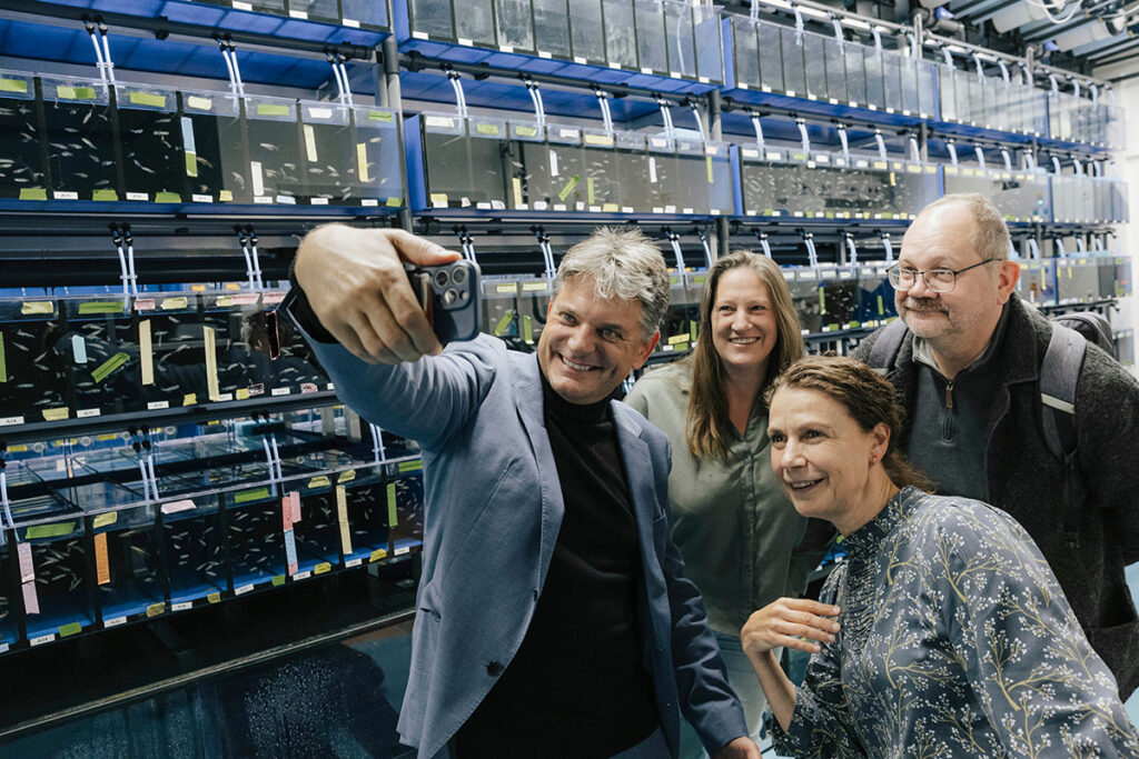 Der Präsident der Universität, Professorin Ober, sowie zwei ihrer Mitarbeiter*innen posieren für ein Selfie. Professor Hornegger hält dafür sein Smartphone hoch, alle schauen in die Kamera und lächeln. Im Hintergrund sind mehrere, wandhohe Reihen mit Fisch-Embryonen aufgestellt.