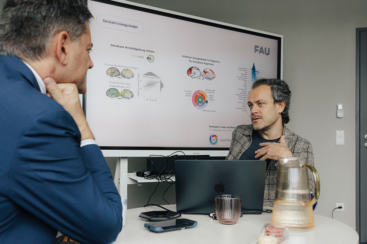 President Hornegger and Prof. Riedl sit on a desk in an office. Hornegger's back is turned to the camera, he is looking at Riedl, who is facing the camera. Behind Riedl is a presentation, with it, he explains something to Hornegger.