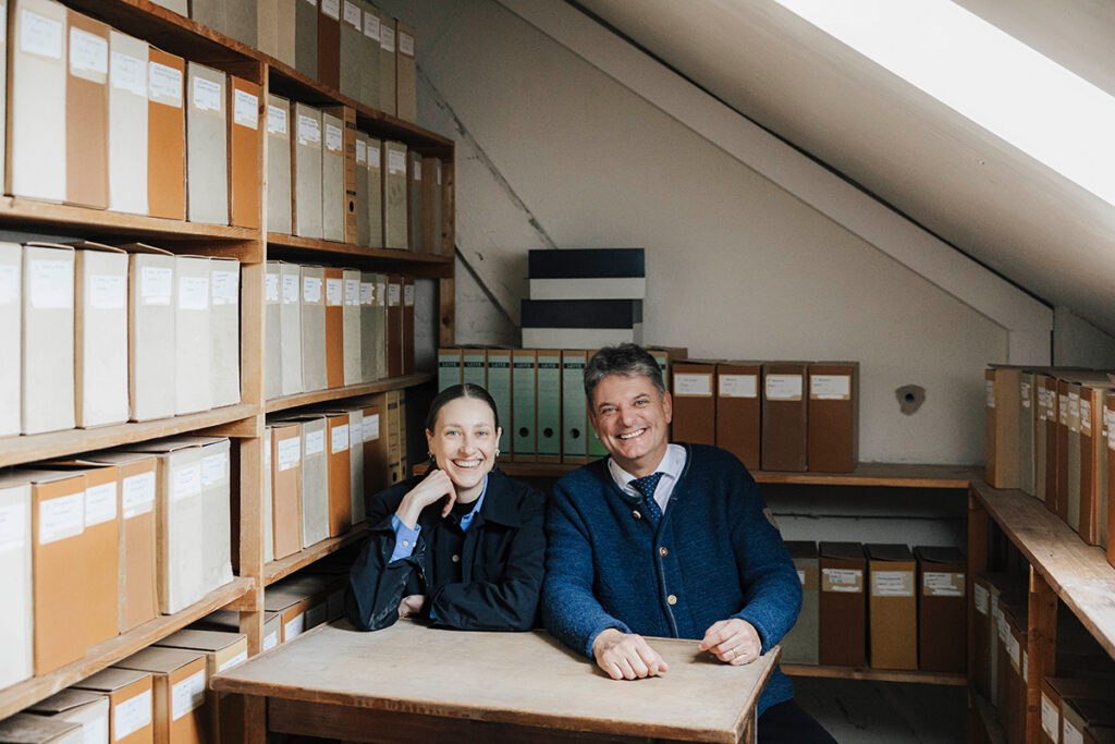 Prof. Pesch und Joachim Hornegger sitzen nebeneinander an einem Tisch und lächeln. Links von ihnen steht eine Bücherwand.