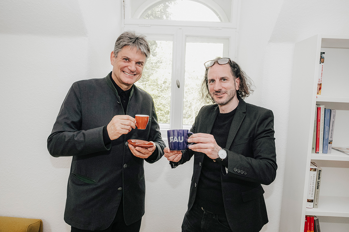 Hornegger and Maltese standing next to each other, smiling at the camera. Hornegger is holding an espresso cup with a saucer, and Malteser an FAU mug.