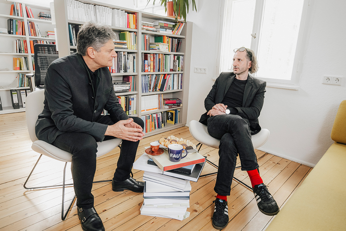 Hornegger and Maltese sitting in a bright room with several large bookcases in the background. In the middle is a pile of books that is acting as a coffee table, with cups and biscuits on it.
