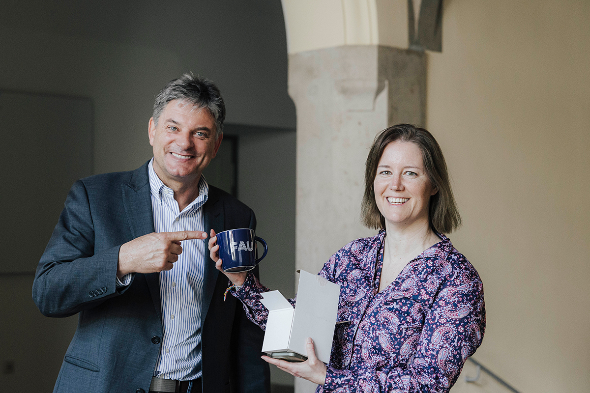 Schilling-Vacaflor and Hornegger are standing together in a corridor holding a cup with an FAU logo.